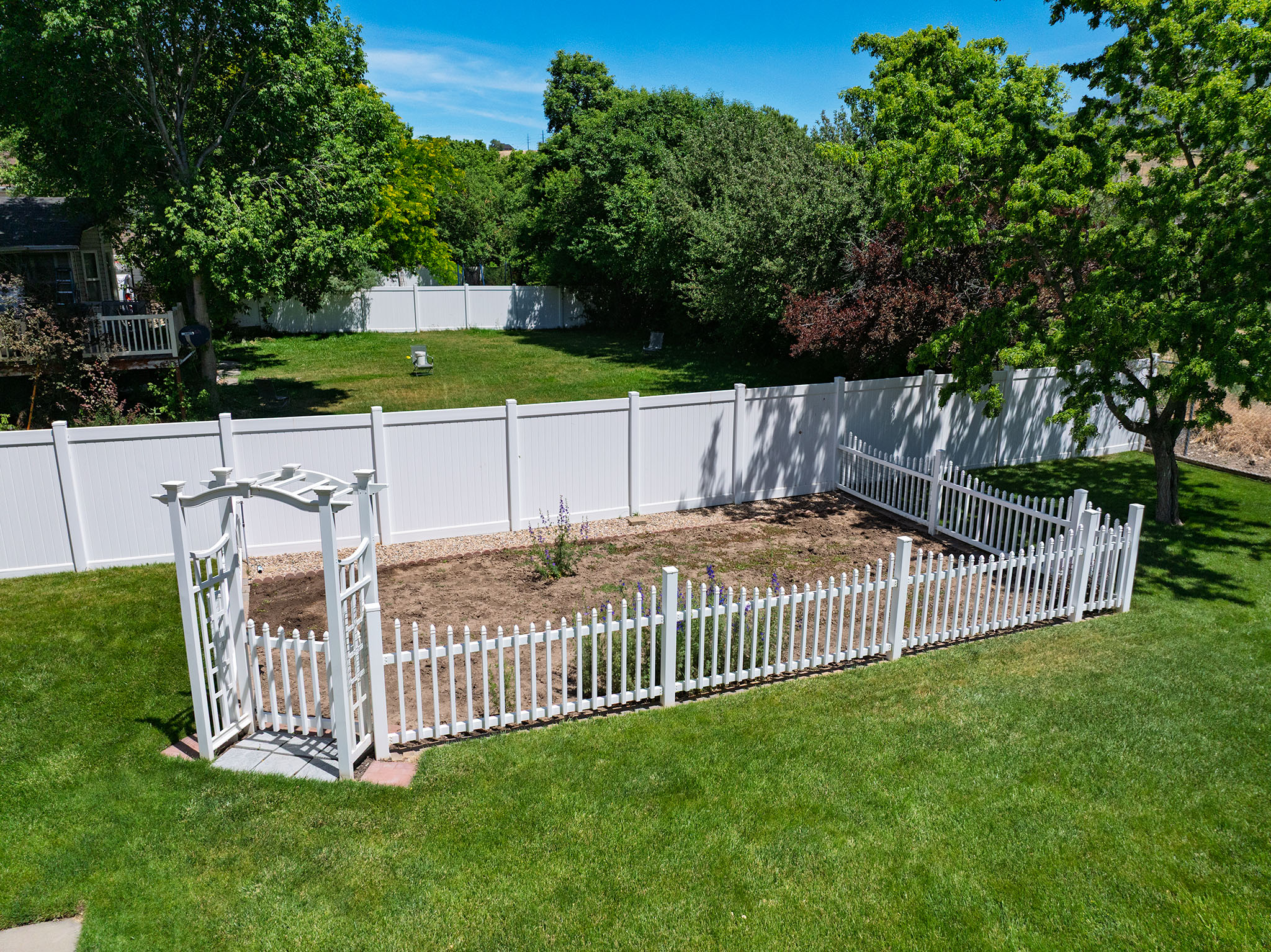Expansive backyard featuring a manicured lawn, white privacy fencing, a decorative white arbor, and a fenced planting area