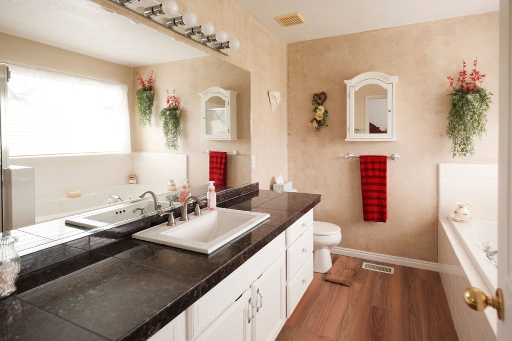 Bathroom featuring a double vanity with dark countertops, white cabinetry, integrated sinks, a built-in bathtub, and wood-finish flooring