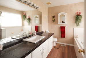 Bathroom featuring a double vanity with dark countertops, white cabinetry, integrated sinks, a built-in bathtub, and wood-finish flooring