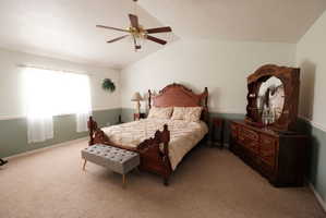 Spacious bedroom featuring vaulted ceilings, a ceiling fan with light fixture, carpeted flooring, and a large window