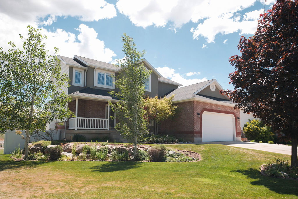 Two-story residence featuring a brick and siding exterior, a covered front porch with white railings, a two-car garage, and mature landscaping