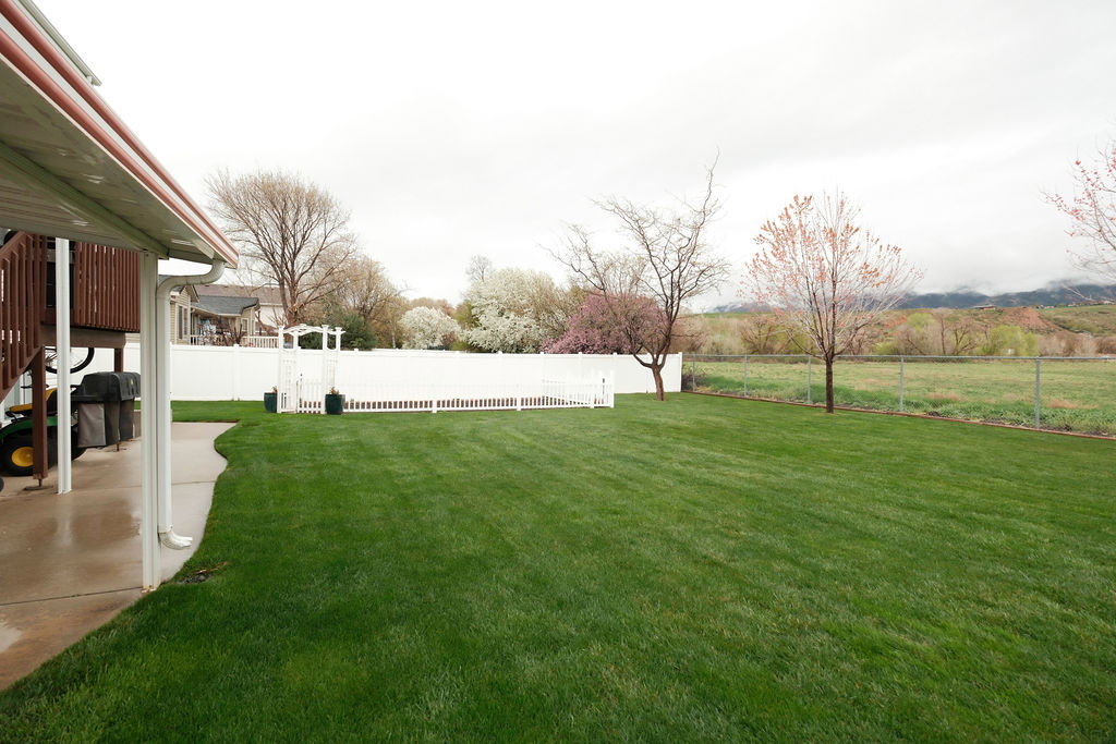 Expansive backyard featuring a large green lawn, white privacy fencing, a decorative white arbor, and a paved patio area under a covered deck