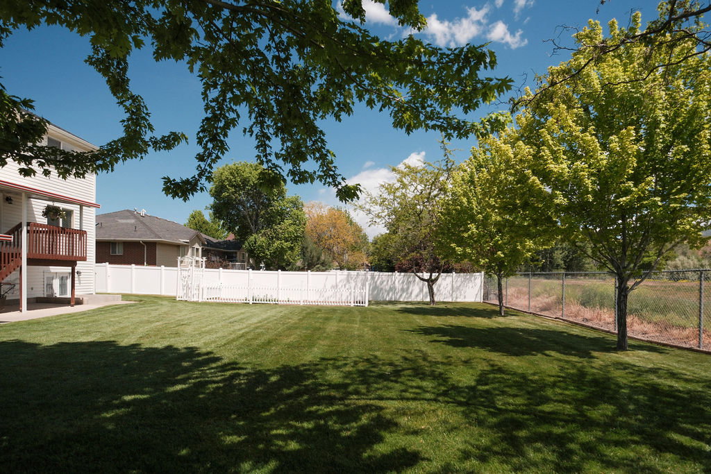 Expansive rear yard featuring a manicured lawn, a white privacy fence, and mature trees