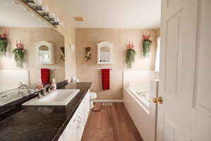 Bathroom featuring an integrated drop-in sink with a brushed nickel faucet, a dark countertop, white base cabinetry, wood-finish flooring, and a built-in tub