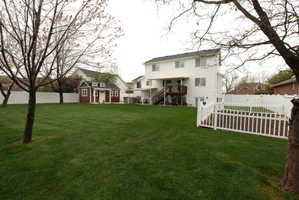 Expansive green lawn featuring a white vinyl privacy fence, a charming red shed with dormer windows, a two-story white exterior residence, and a raised wooden deck with stairs