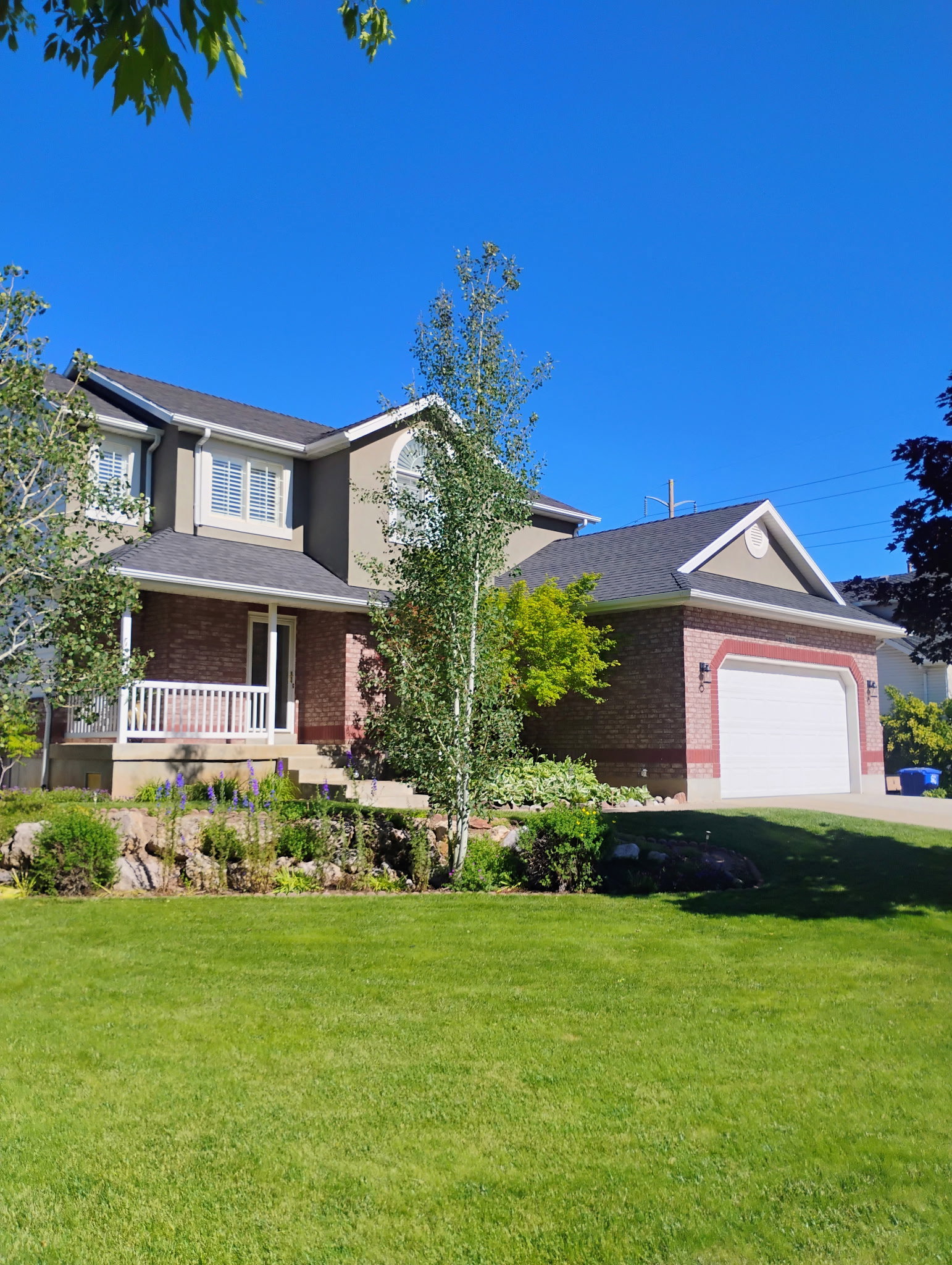 Two-story residence featuring a brick and stucco exterior, an attached two-car garage with an arched trim, a covered front porch with white railings, and a landscaped front yard with a manicured lawn