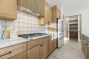 Kitchen featuring decorative backsplash, high end white refrigerator, stainless steel gas cooktop, light stone countertops, and light floors