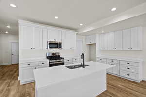 Kitchen featuring stainless steel appliances, a center island with sink, white cabinetry, light stone countertops, and light wood-style floors