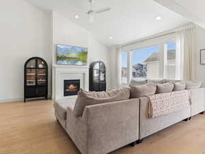 Living room with light wood-type flooring, ceiling fan, a lit fireplace, lofted ceiling, and recessed lighting