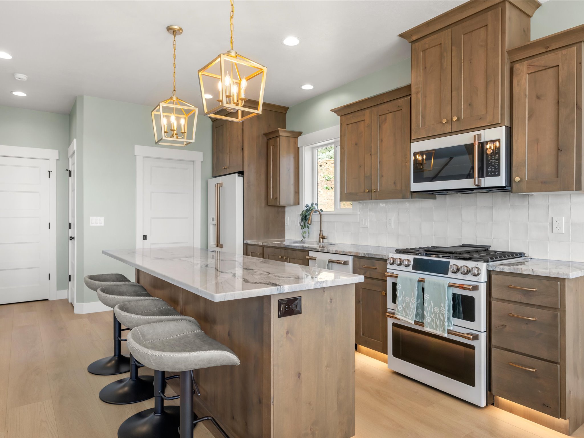 Modern kitchen featuring a large island with a light-toned countertop, wood-finish cabinetry, stainless steel appliances, a tile backsplash, and two geometric pendant lights