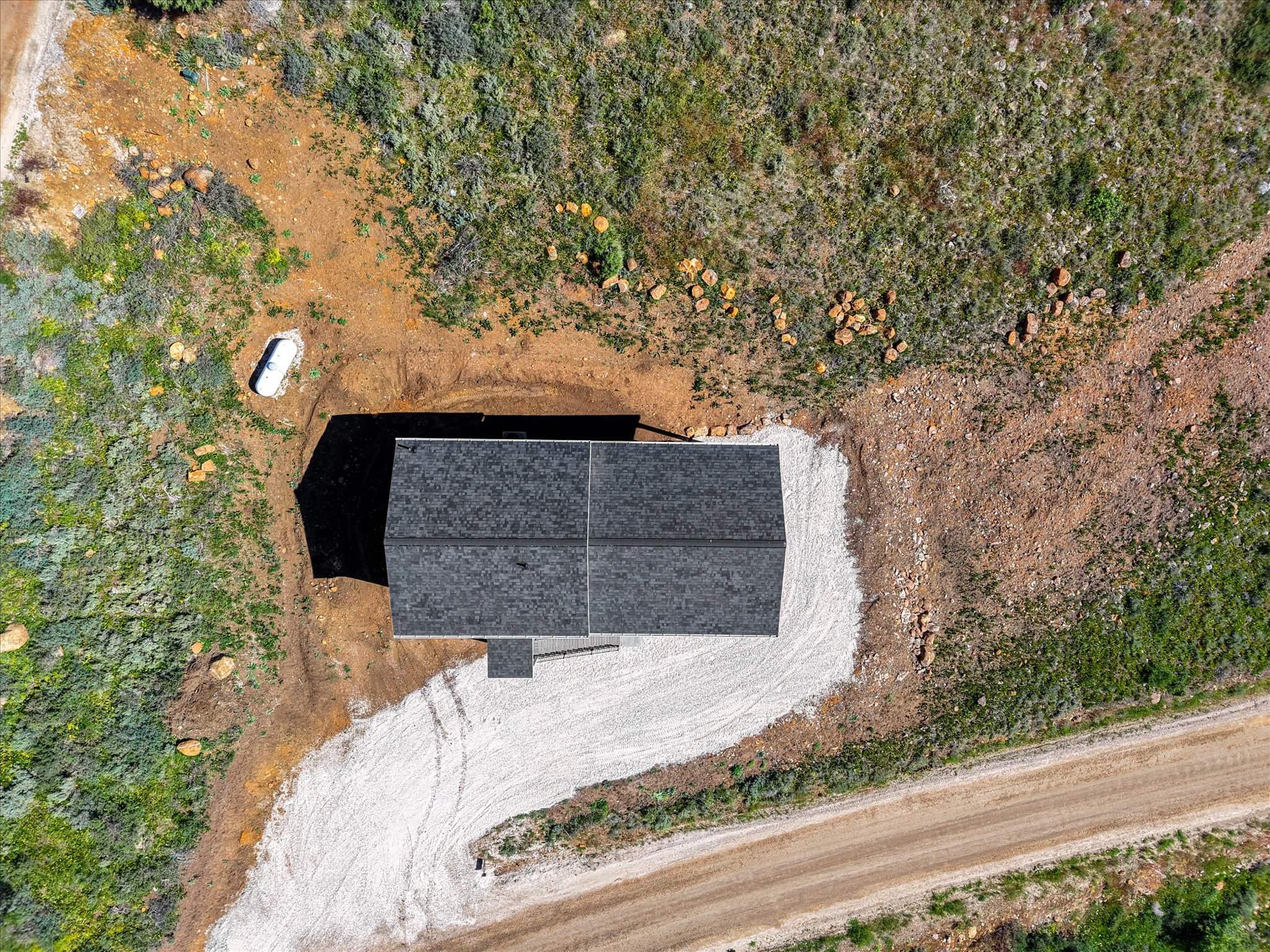 New construction featuring a dark shingle roof, surrounded by natural terrain and a gravel driveway