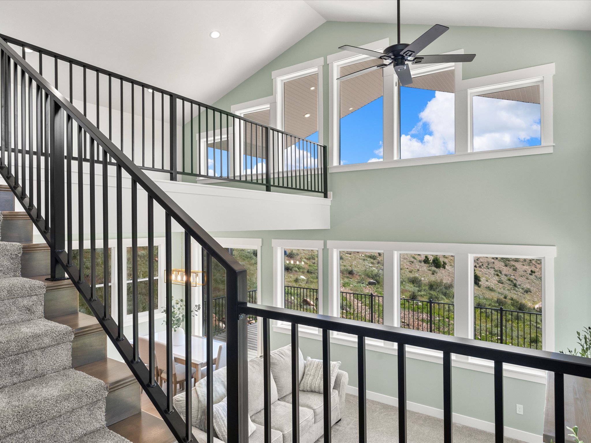 Two-story living area featuring a vaulted ceiling with a ceiling fan, multiple large windows, and a carpeted staircase with dark metal railings
