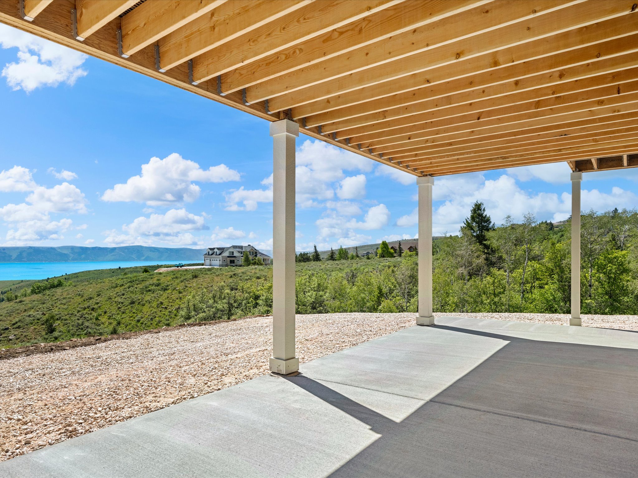 Expansive concrete patio with overhead wood joist structure, supported by square columns, overlooking a distant water body and rolling hills