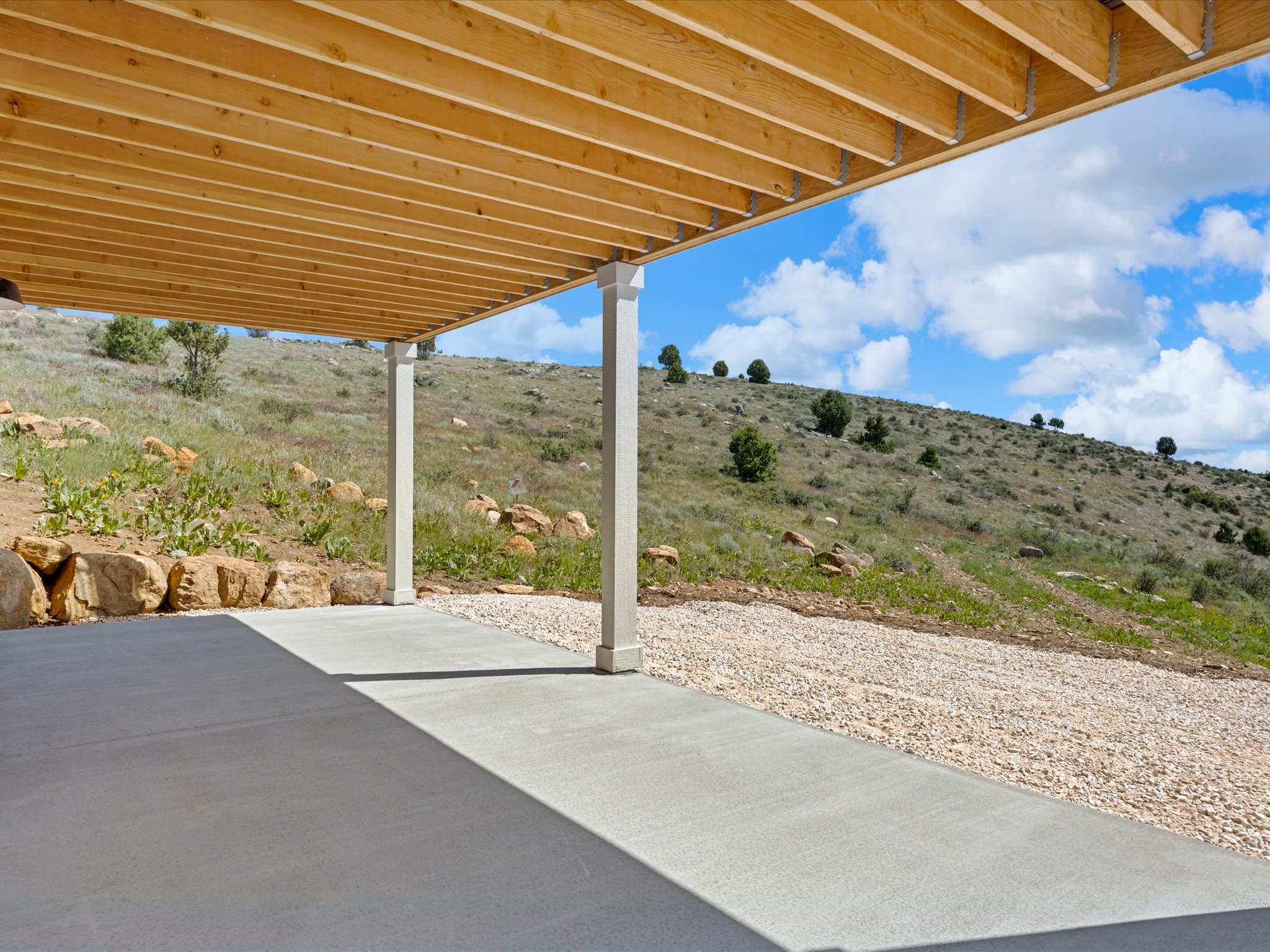Covered concrete patio with two support columns, overlooking a sloped natural landscape with scattered trees and rock formations
