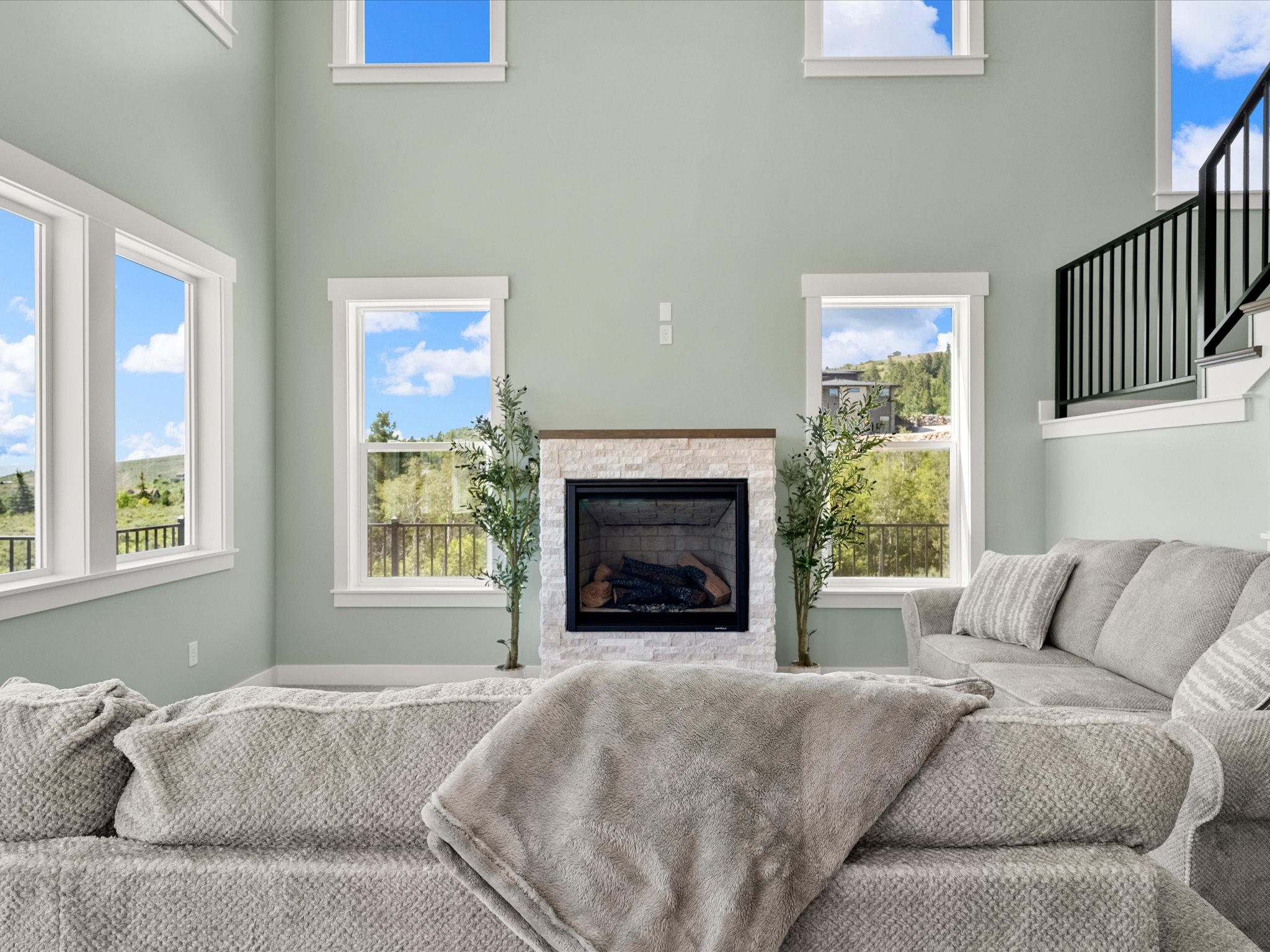 Spacious living area featuring a two-story ceiling, multiple windows, a stone-clad fireplace with a wood mantel, and a staircase with black metal balusters