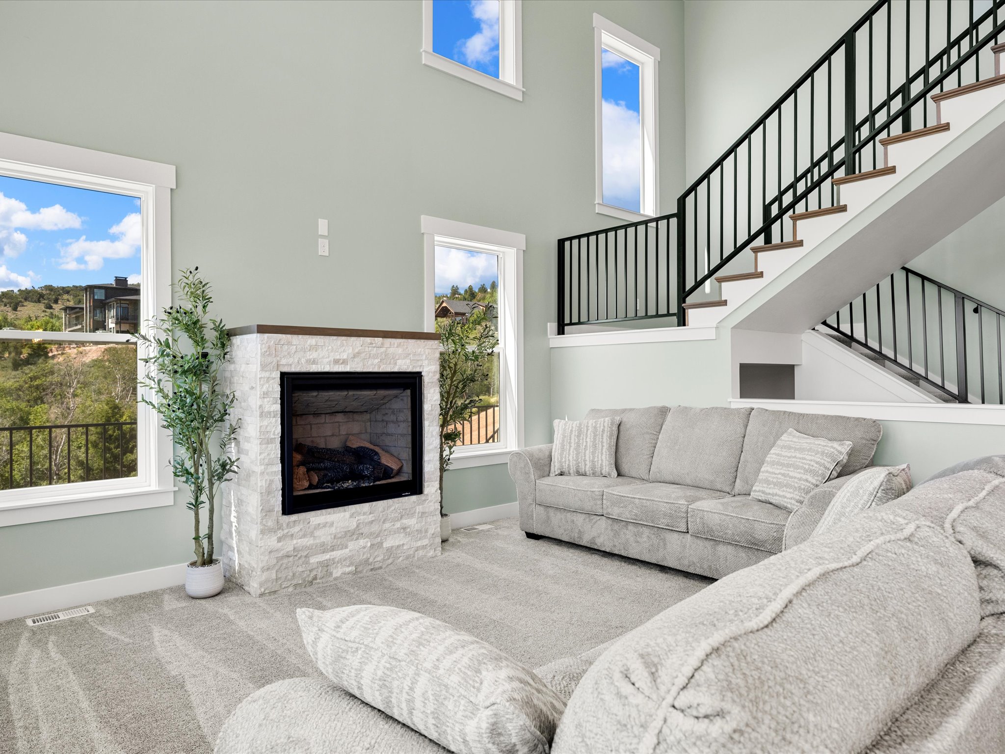 Two-story living area featuring a stacked stone fireplace with a dark wood mantel, multiple windows, and a staircase with open risers and black metal railings