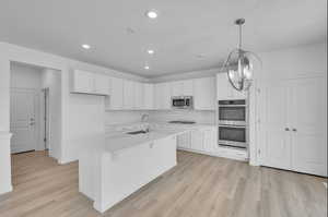 Kitchen with white cabinetry, stainless steel appliances, light wood-style flooring, and a center island with sink