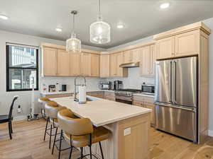 Kitchen with light wood finish cabinetry, high quality appliances, an island with sink, hanging light fixtures, and light wood-type flooring