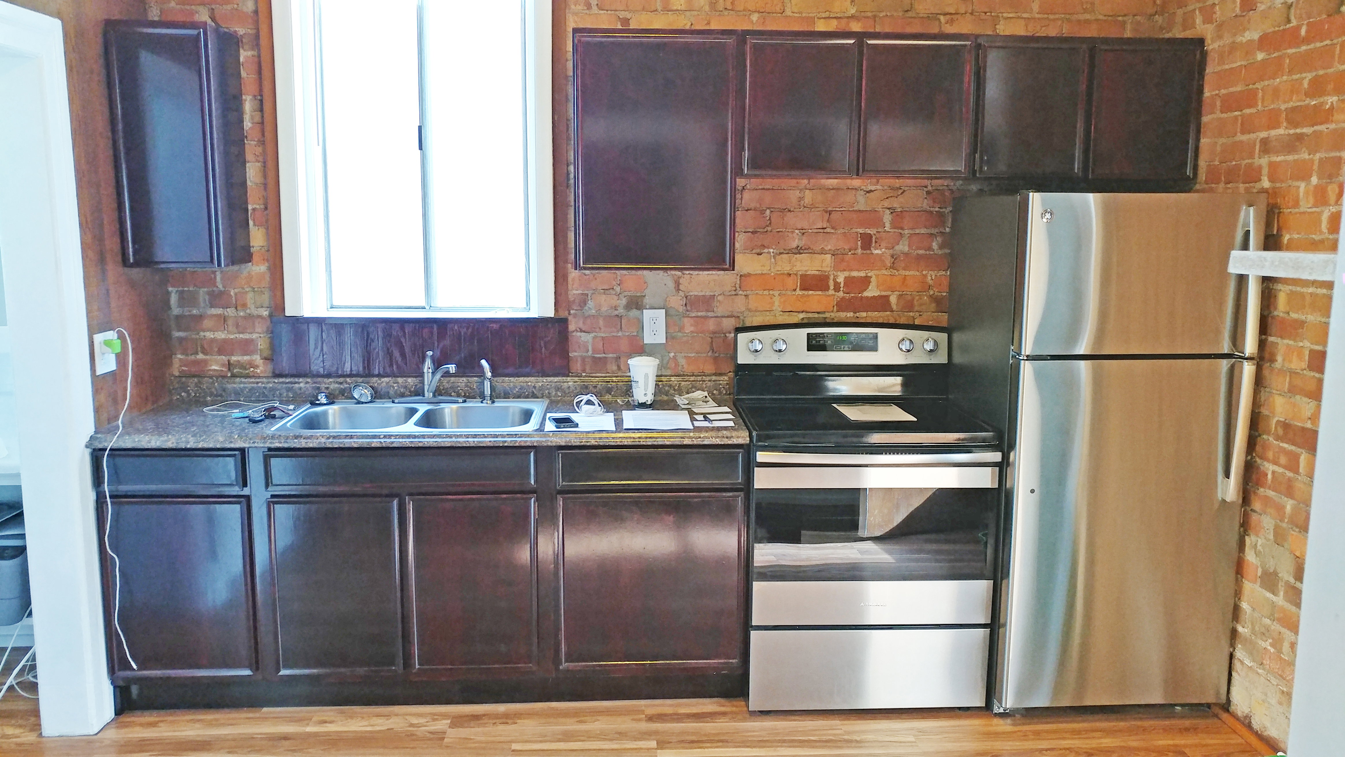 Kitchen with stainless steel appliances, dark wood finish cabinets, brick wall, and light wood-style floors