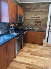 Kitchen featuring wood finish cabinets, stainless steel appliances, dark stone countertops, light wood-style flooring, and brick wall