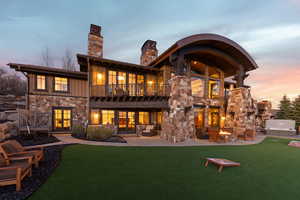 Back of house at dusk with stone siding, a balcony, board and batten siding, a lawn, and a patio area