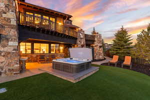 Back of property at dusk with a balcony, an outdoor saltwater hot tub, stone siding, a yard, and a patio area