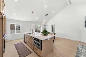 Kitchen with vaulted ceiling, light stone counters, an island with sink, decorative light fixtures, and light wood-style floors