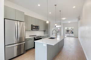 Kitchen featuring stainless steel appliances, gray cabinets, a kitchen island with sink, decorative light fixtures, and light wood-type flooring
