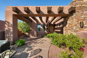 Entryway featuring an architectural pergola with exposed timber beams, stucco exterior, stacked stone wall accents, a paved walkway, and low-maintenance landscaping