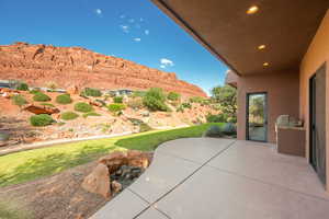 Covered patio with integrated outdoor grill station and recessed lighting, overlooking a landscaped yard with natural rock formations and distant red rock cliffs