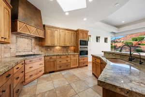 Spacious kitchen featuring custom wood cabinetry, granite countertops, tile backsplash, stainless steel built-in appliances, and a central island with an undermount sink