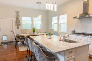 Kitchen with white cabinetry, decorative light fixtures, a center island with sink, light stone counters, and dark wood-style flooring