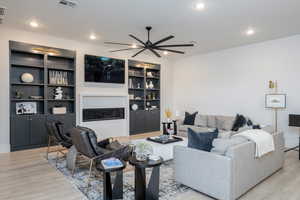 Living room featuring light wood-style flooring, a ceiling fan, built in shelves, a glass covered fireplace, and recessed lighting