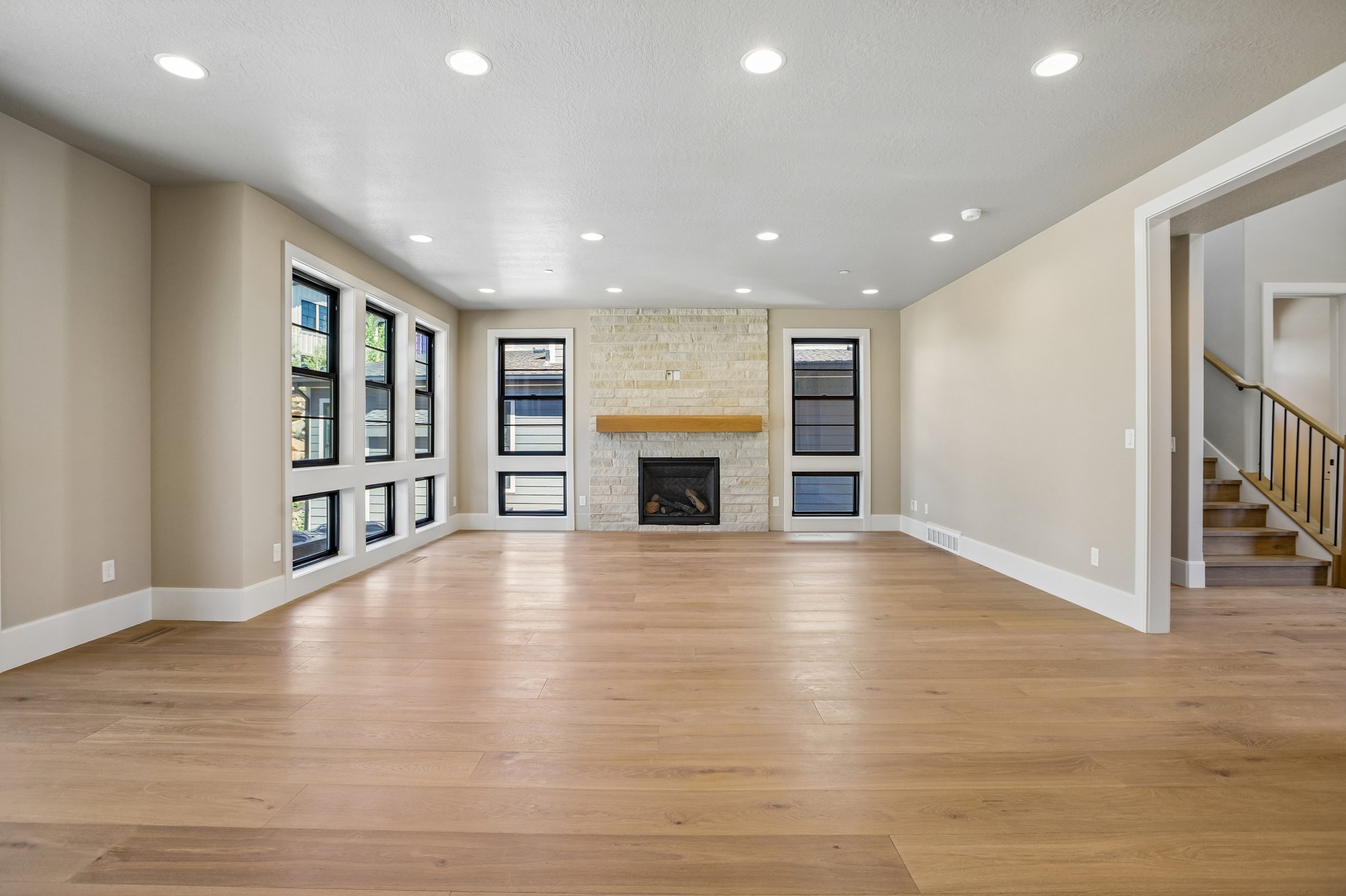 Spacious living area featuring light wood-finish flooring, a stone-clad fireplace with a wood mantle, recessed lighting, and multiple windows with black frames