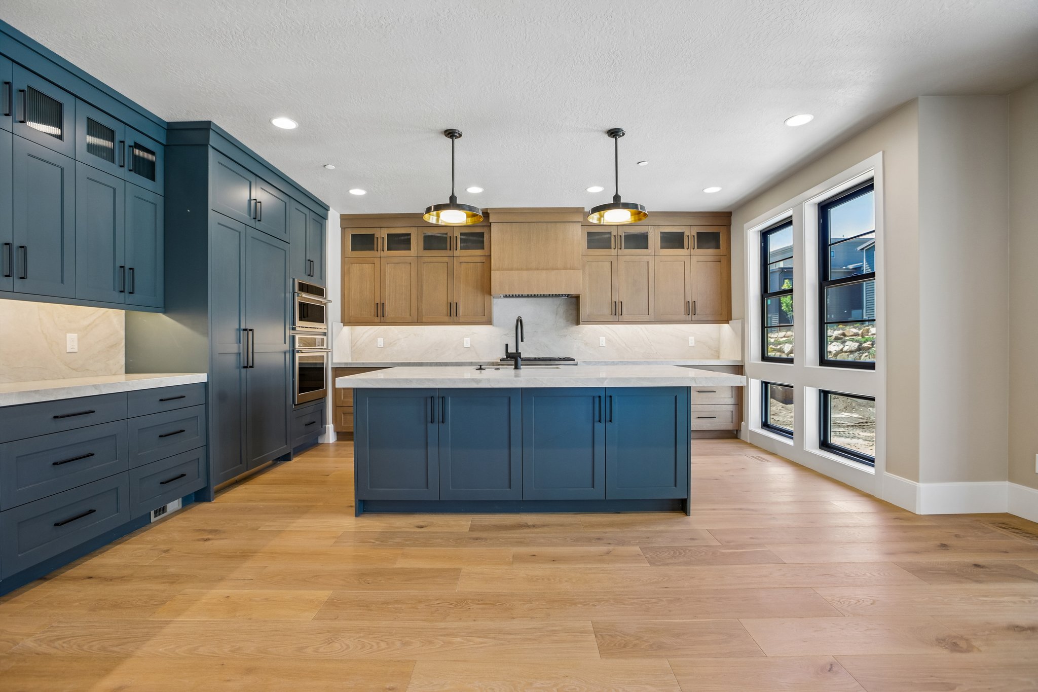 Open-concept kitchen featuring an expansive island with a waterfall countertop, dual-tone cabinetry, wood-finish flooring, built-in appliances, and large windows with black frames