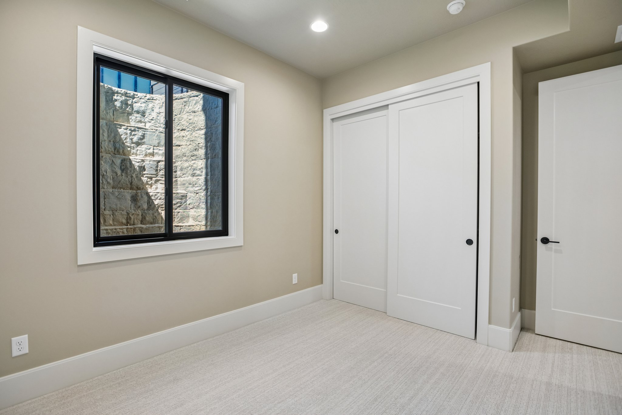 Recessed lighting illuminates a neutral-toned room featuring a window with black trim, white sliding closet doors, and light-colored carpeting