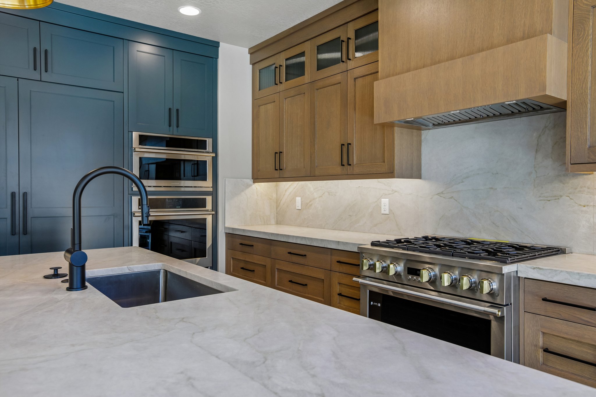 Kitchen island featuring an undermount sink with a matte black high-arc faucet, light-toned countertops, and integrated stainless steel double ovens
