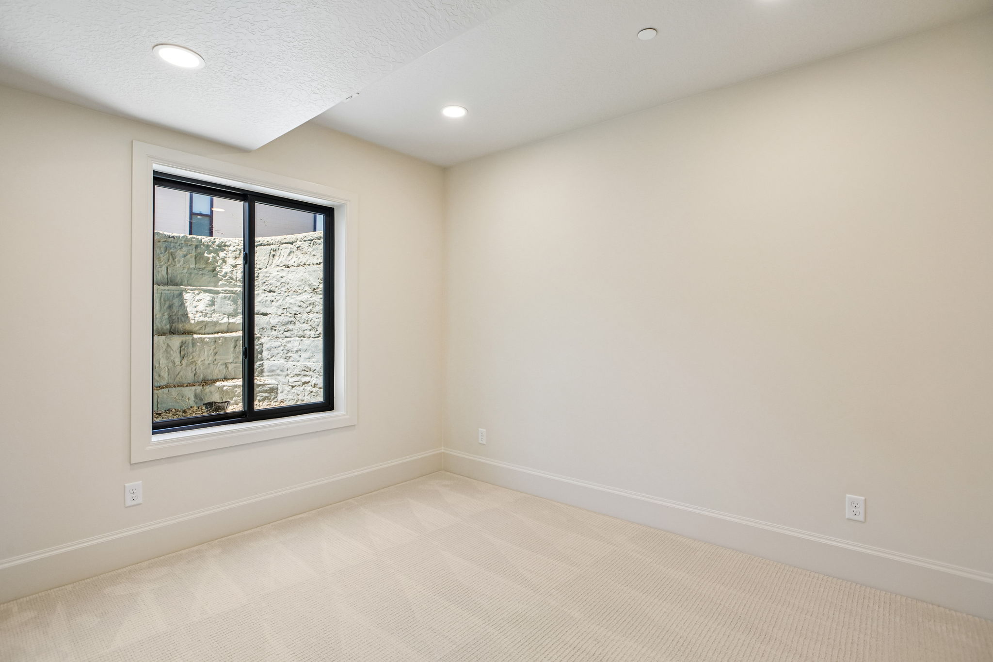 Room featuring neutral wall tones, recessed lighting, white baseboards, and a large window with black framing overlooking a stone retaining wall