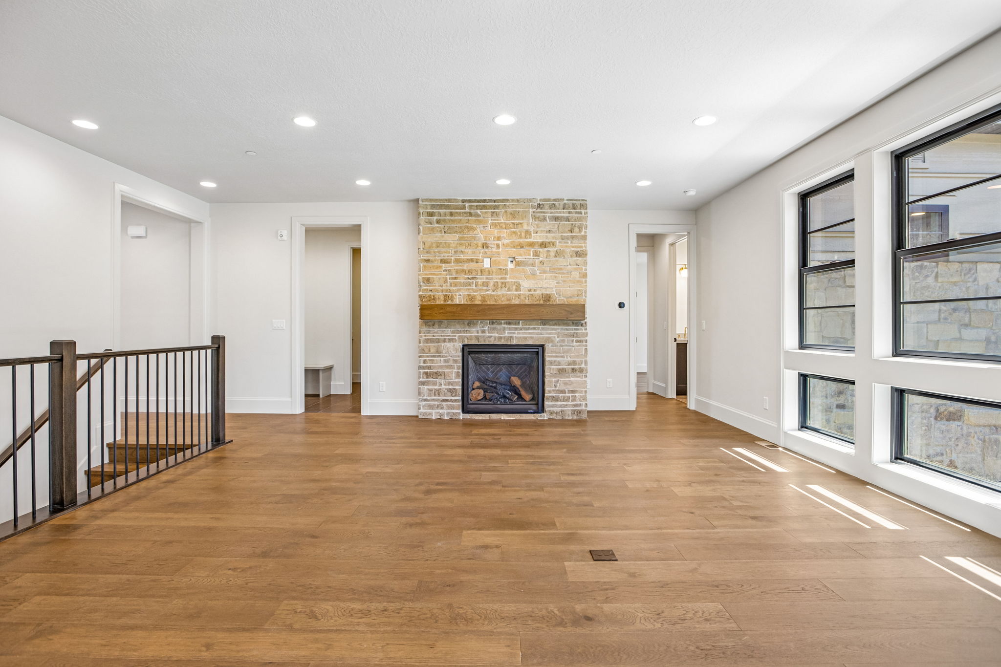 Spacious living area featuring a stacked stone fireplace with a substantial wood mantel, extensive wood-finish flooring, recessed lighting, large multi-pane windows, and a staircase with a dark wood handrail and black metal balusters