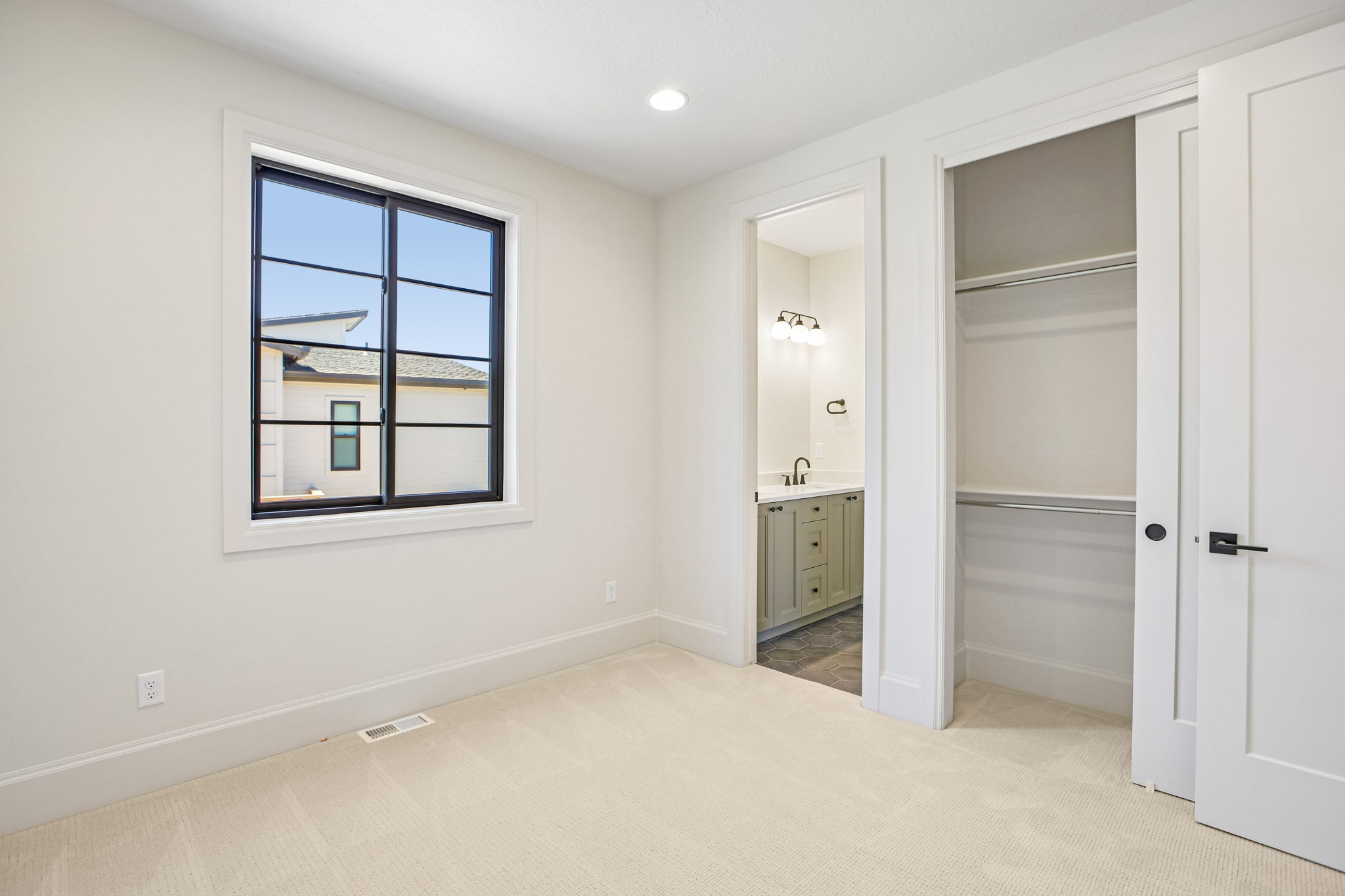 Bright room featuring a grid-pattern window with black framing, neutral carpet flooring, an integrated closet with shelving and hanging rods, and a doorway to a bathroom with a sage green vanity and wall-mounted light fixture