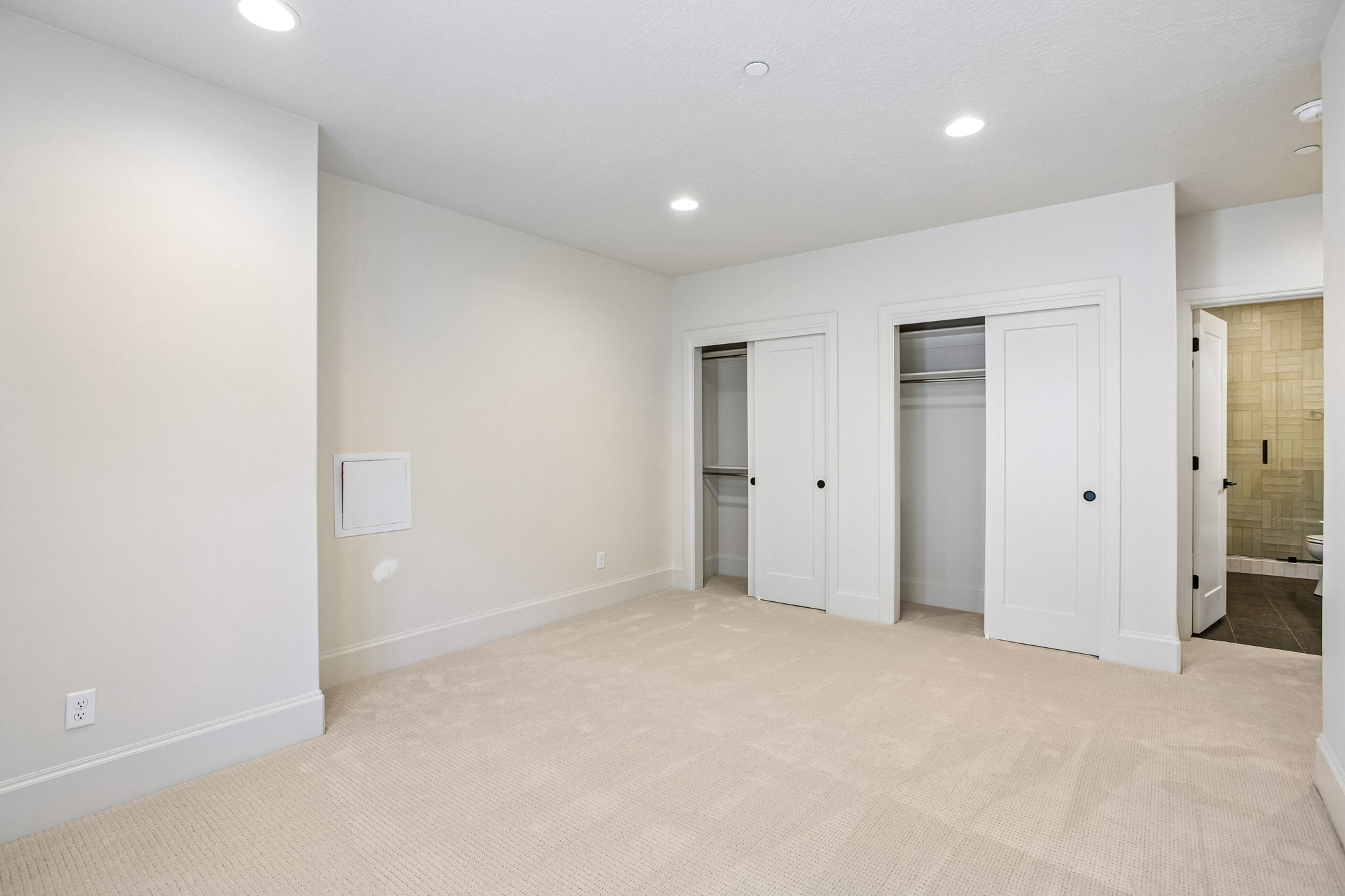 Neutral-toned room with plush carpet flooring, recessed lighting, two sliding closet doors with black hardware, and an open doorway to a tiled bathroom