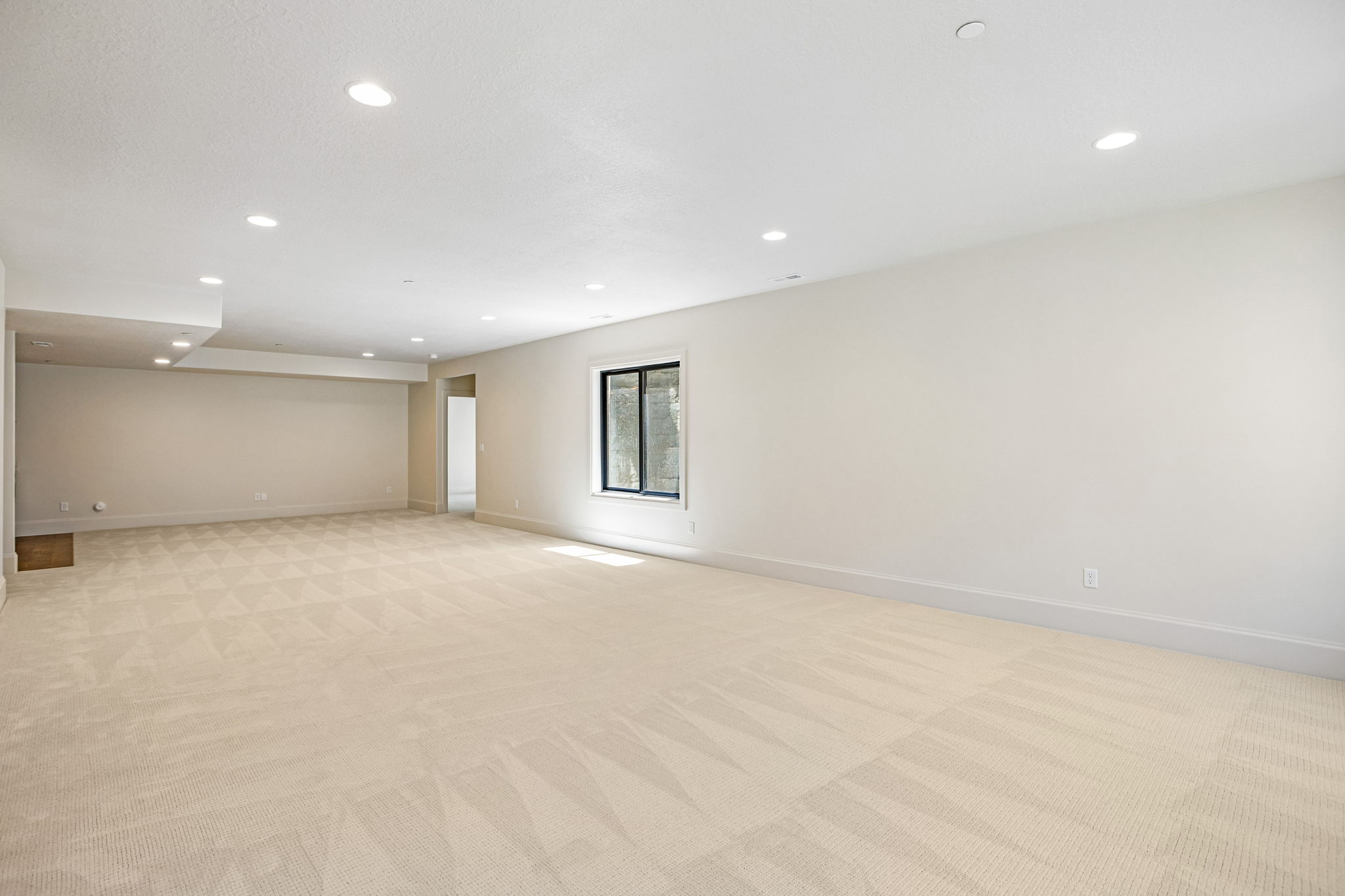 Spacious interior room featuring light-toned carpeting with a subtle geometric pattern, recessed lighting, and a large window with black framing