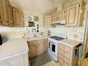Kitchen featuring white gas range oven, a textured ceiling, light countertops, wallpapered walls, and light wood finish cabinets