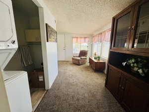 Laundry area with a textured ceiling, light colored carpet, and stacked washing machine and dryer
