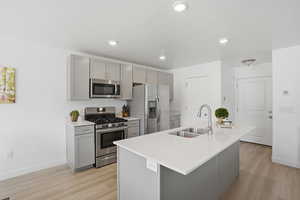Kitchen with gray cabinetry, stainless steel appliances, a kitchen island with sink, light flooring, and light stone counters