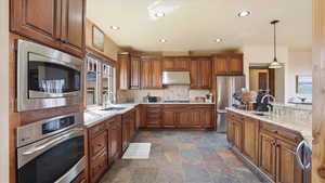 Kitchen featuring stainless steel appliances, wood finish cabinetry, hanging light fixtures, stone finish flooring, and light stone counters