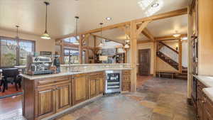 Kitchen with beverage cooler, wood finish cabinetry, stone tile floors, light stone countertops, and suspended lighting