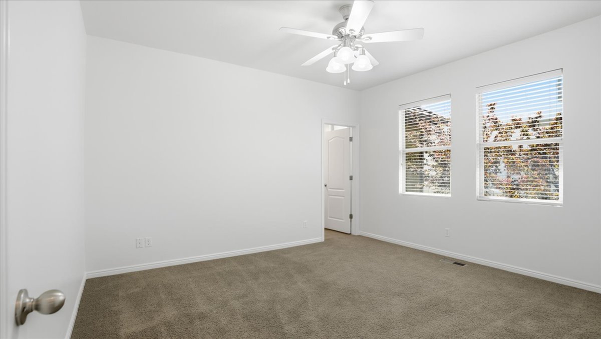 Spacious room featuring neutral carpeting, white painted walls, two windows with blinds, a white ceiling fan with light fixture, and a white interior door