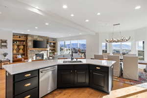 Kitchen featuring dark cabinetry, light wood finished floors, a stone fireplace, dishwasher, and a kitchen island with sink