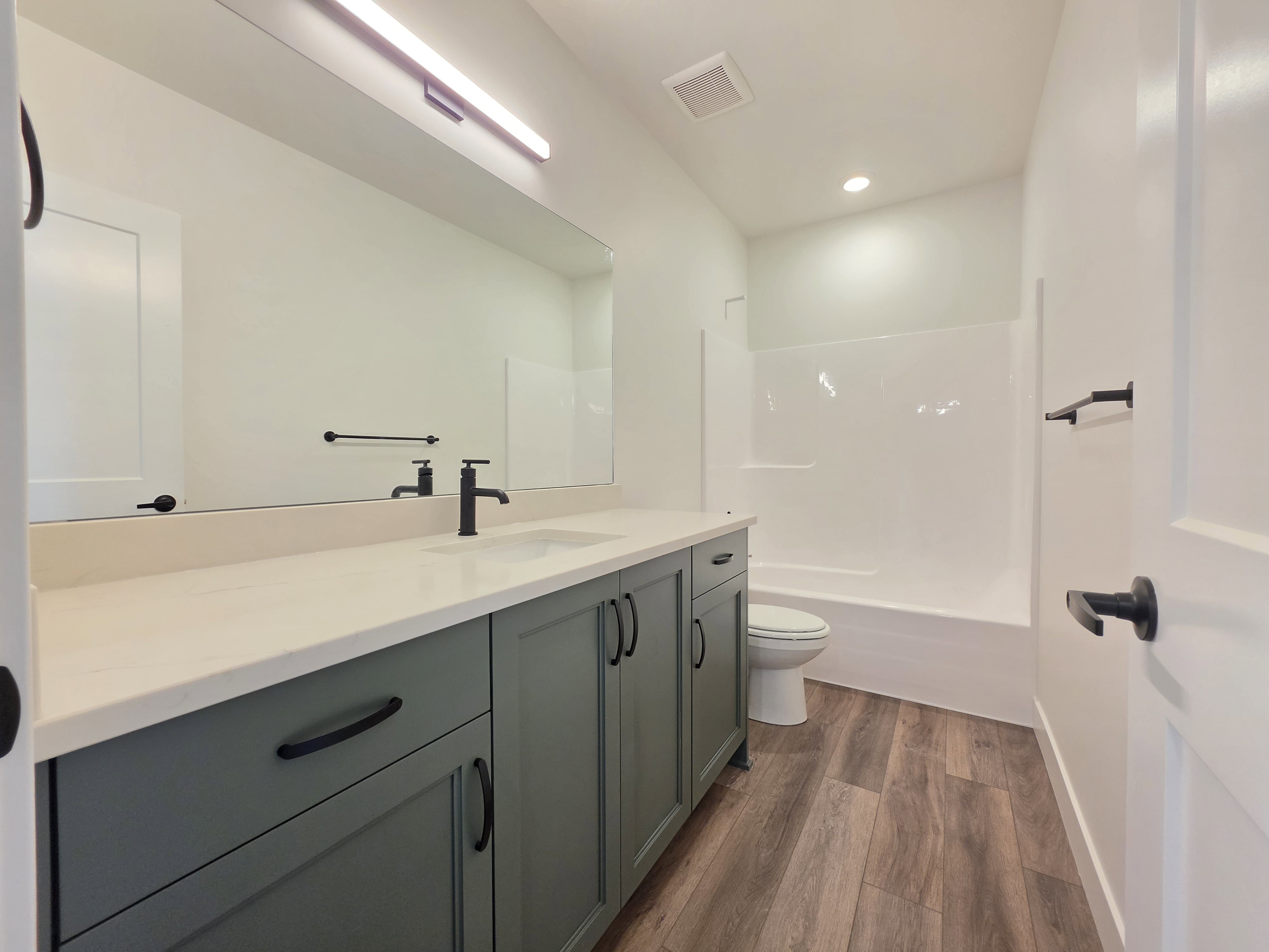 Bathroom featuring a green vanity with a light countertop, wood-finish flooring, a full-width mirror, and a white tub with shower surround